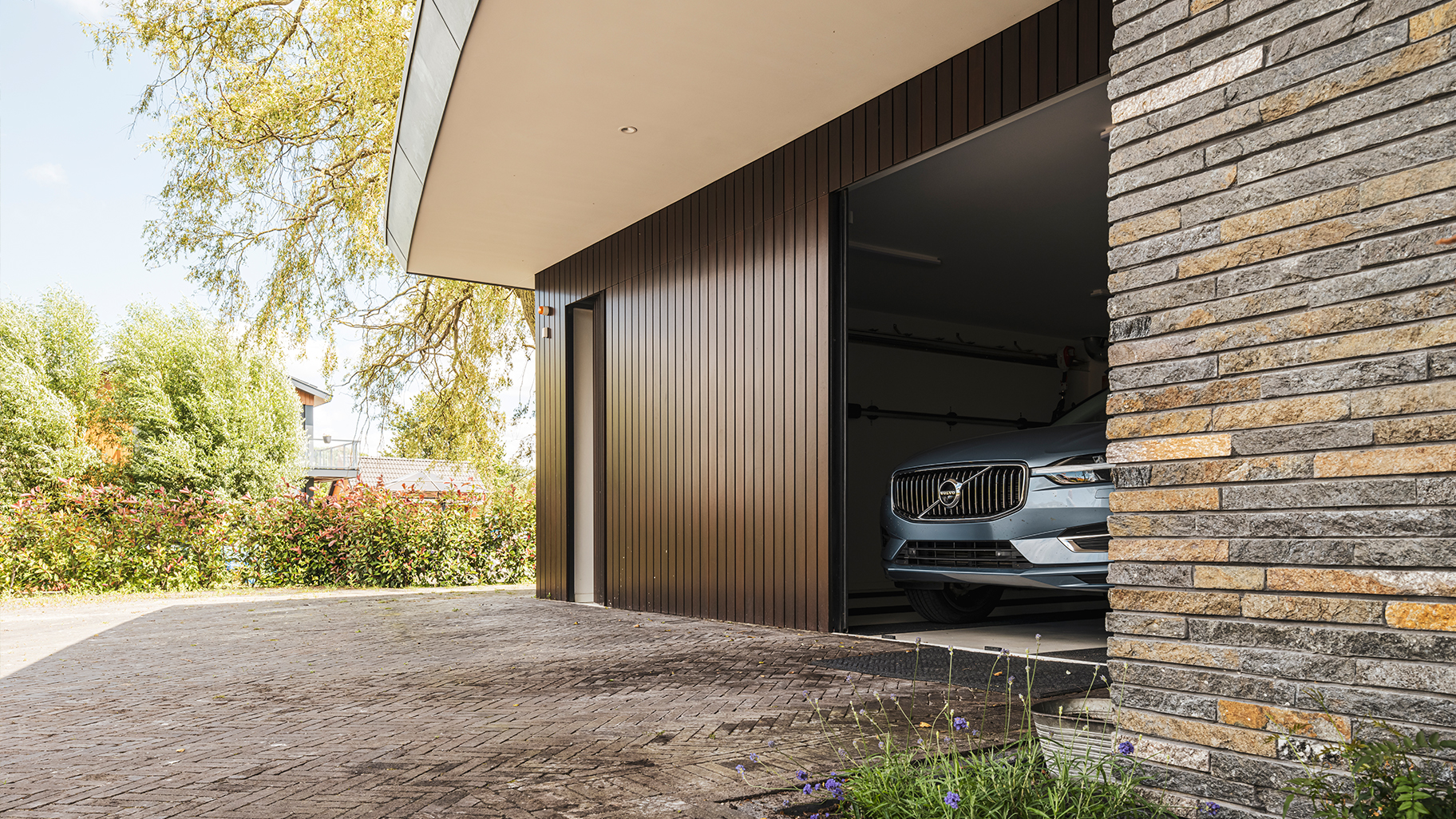 Onzichtbare zijwaartse garagedeur volledig gelijk is met de gevel geplaatst. Hierdoor lijkt de deur, wanneer gesloten, onzichtbaar en vormt deze een perfect doorlopende houten wand. De verticale houten latten geven de gevel een moderne, strakke uitstraling.