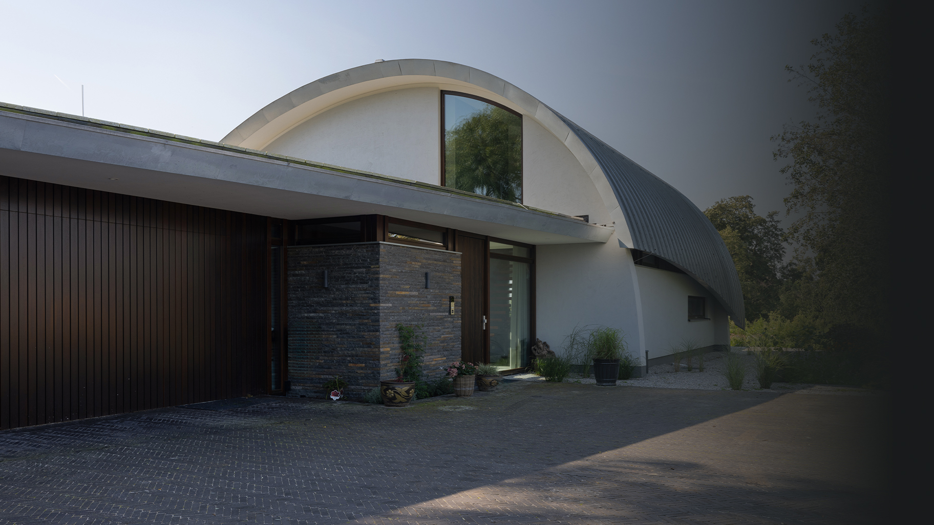 Onzichtbare zijwaartse garagedeur volledig gelijk is met de gevel geplaatst. Hierdoor lijkt de deur, wanneer gesloten, onzichtbaar en vormt deze een perfect doorlopende houten wand. De verticale houten latten geven de gevel een moderne, strakke uitstraling.