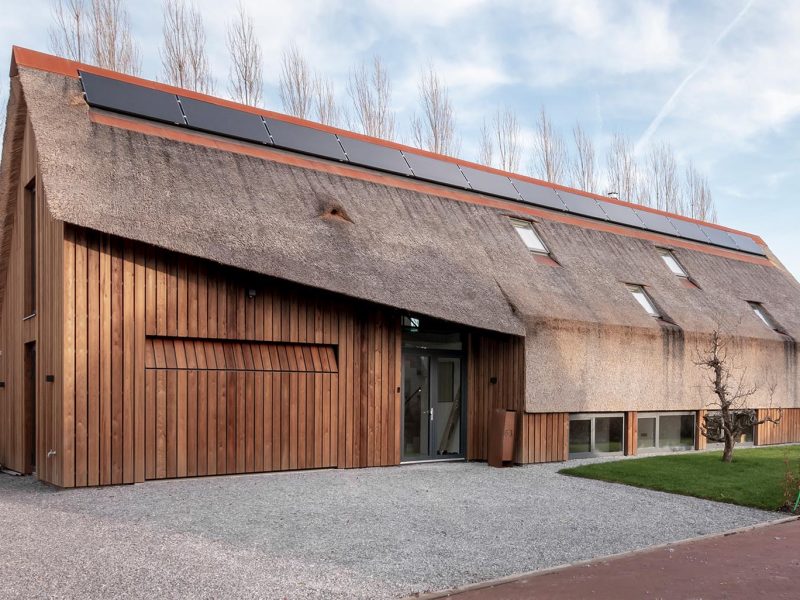 Moderne rietgedekte woning die een gevelrenovatie heeft gehad, met een onopvallende verticale houten garagedeur die volledig opgaat in de gevel en verrast wanneer geopend
