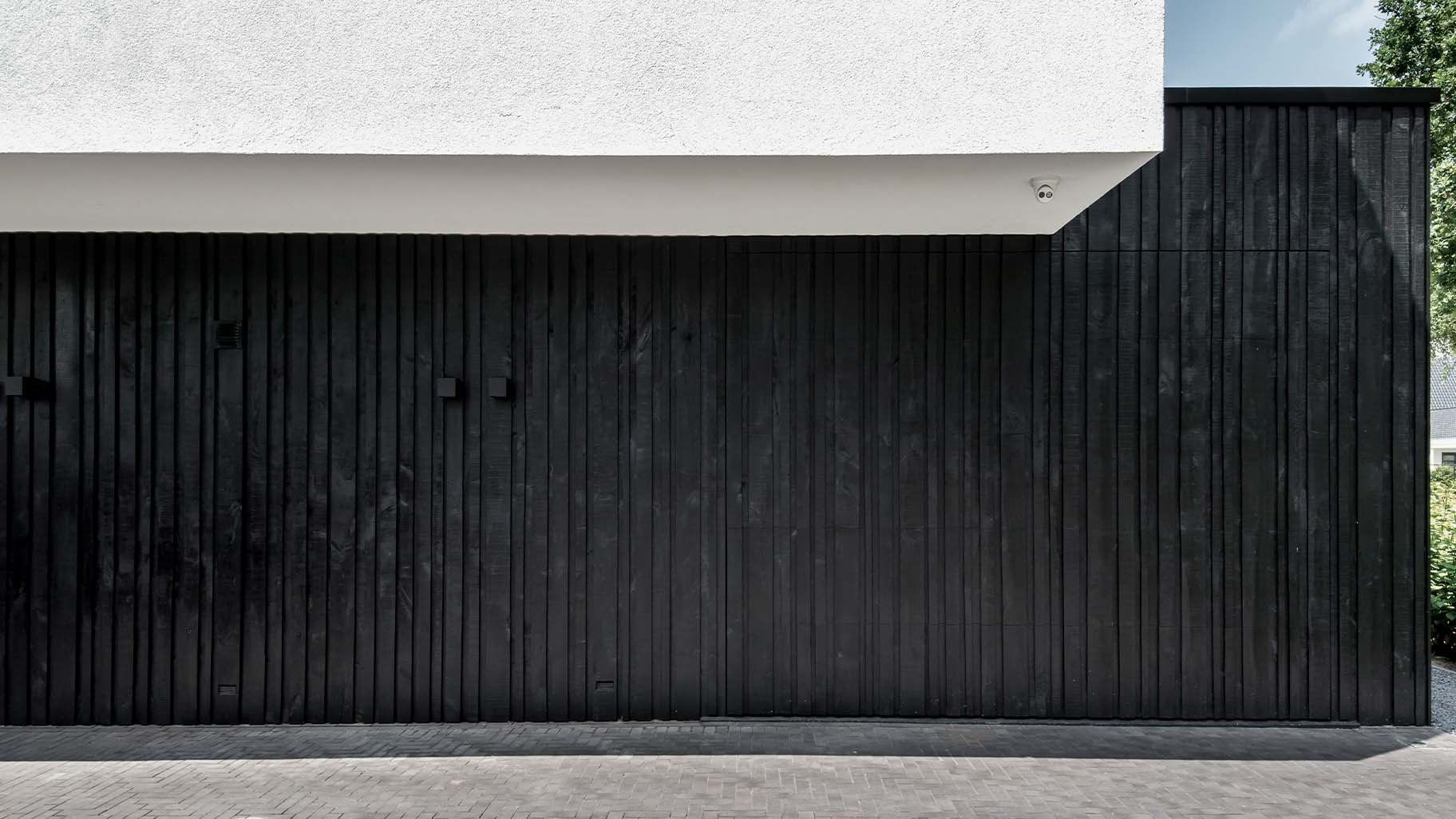 Moderne zwarte garagedeur met verticale houten latten in diverse breedtes, contrasterend met witte gevel
